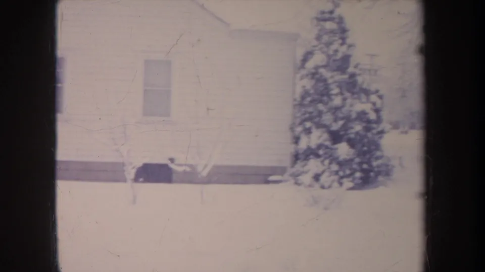 1968 Michigan 8mm frame showing the side of a white house, a snowy evergreen shrub, and a bare sapling in the yard.