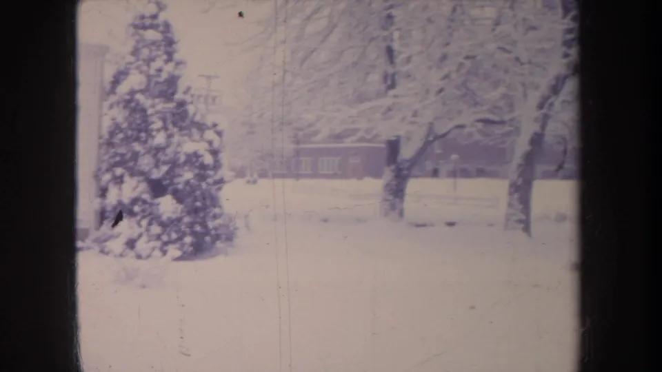 1968 Michigan 8mm frame showing snowy trees and a distant low school or neighborhood building across a blanketed yard.