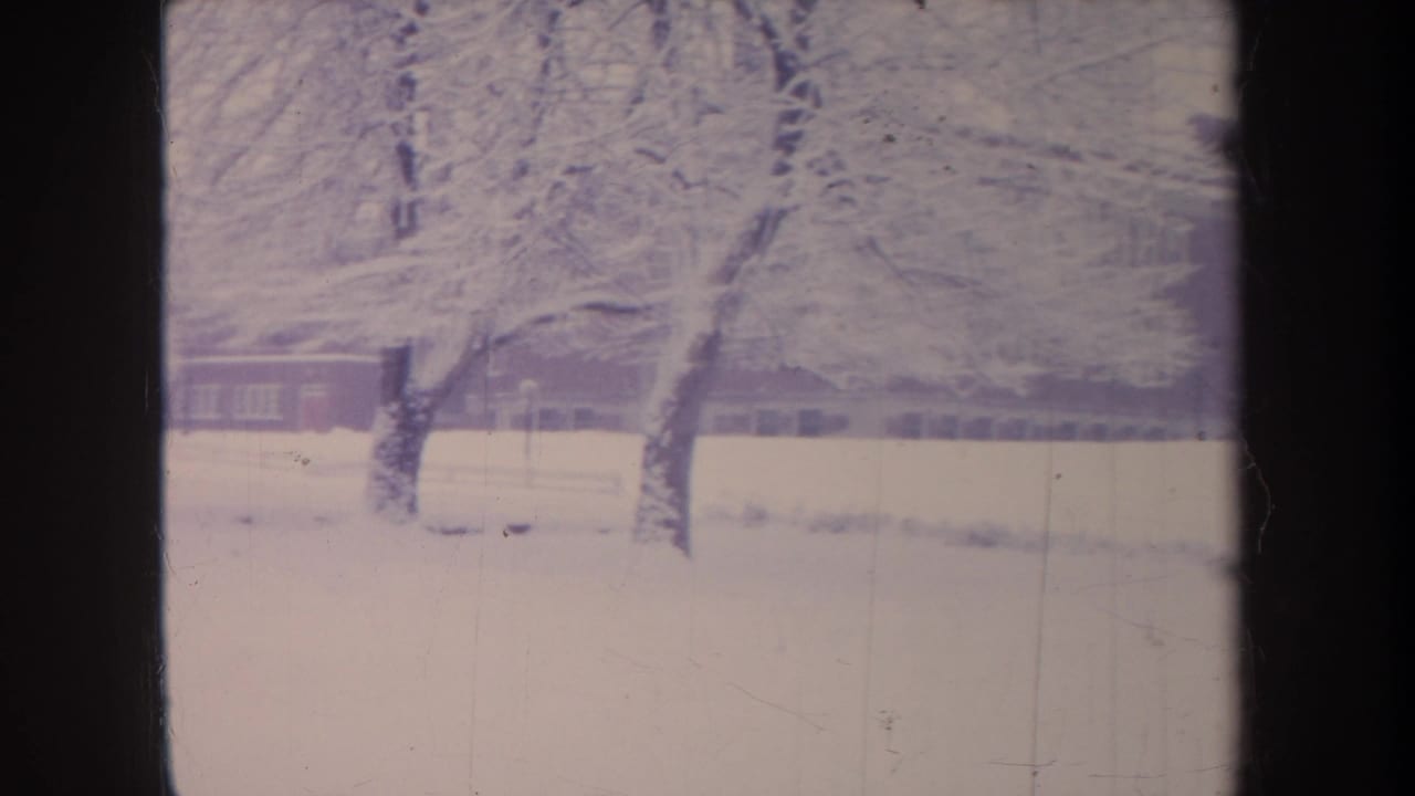 1968 Michigan winter frame with heavy snow on branches and a long brick school-like building beyond the snowy grounds.