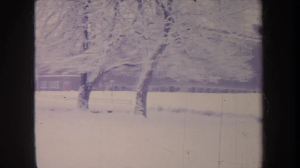 1968 Michigan winter frame with heavy snow on branches and a long brick school-like building beyond the snowy grounds.