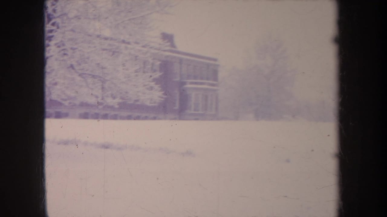8mm home-movie frame from Michigan showing a snow-covered yard and a partial brick school building during winter.