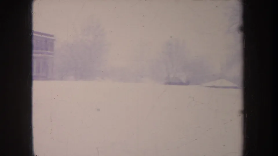 Soft 1968 Michigan winter frame with an open snowfield, distant bare trees, and part of a brick building at the edge.