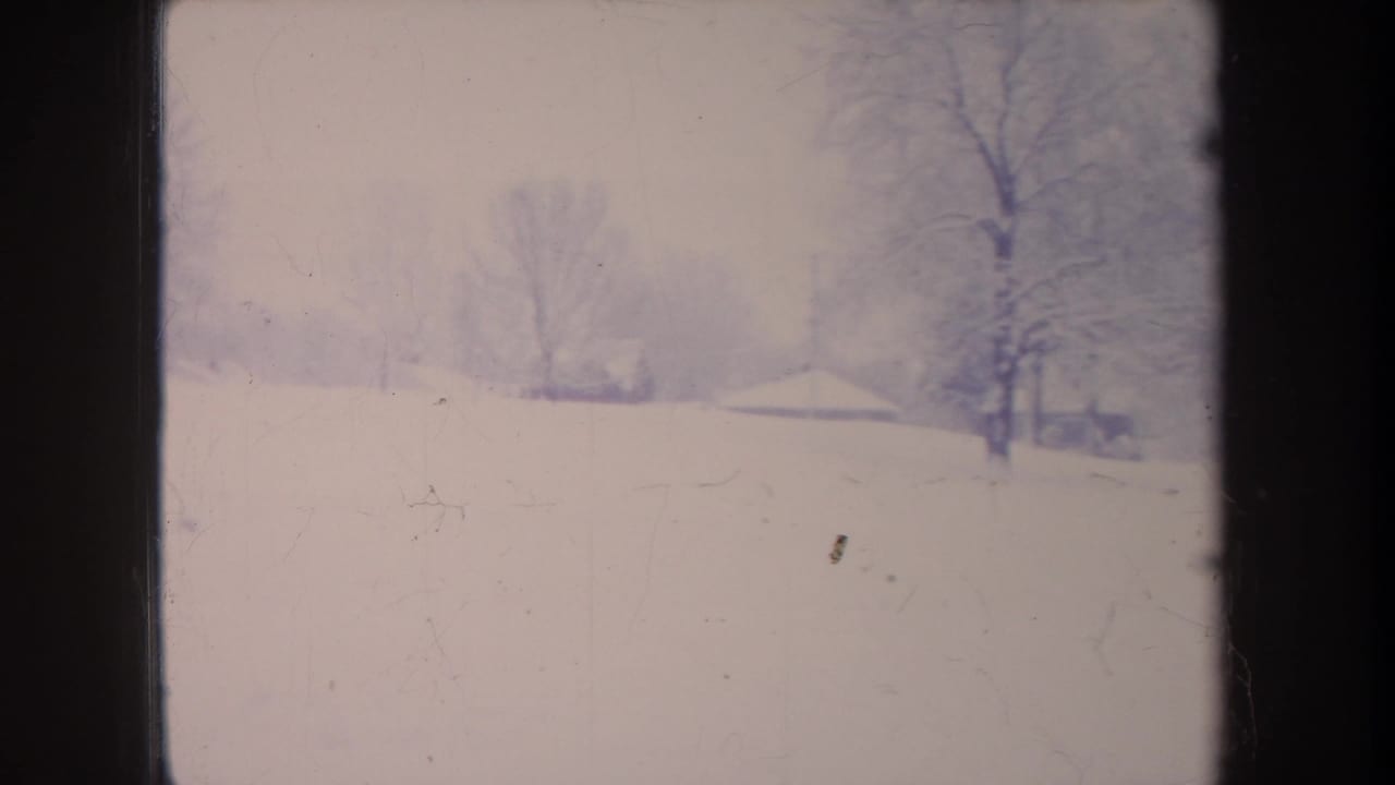 1968 Michigan 8mm frame showing a snow-covered field, leafless trees, and faint distant buildings through pale winter haze.