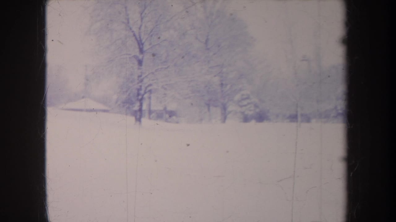 1968 Michigan winter frame with open snow cover, bare tree trunks, and faint houses or structures along the hazy horizon.