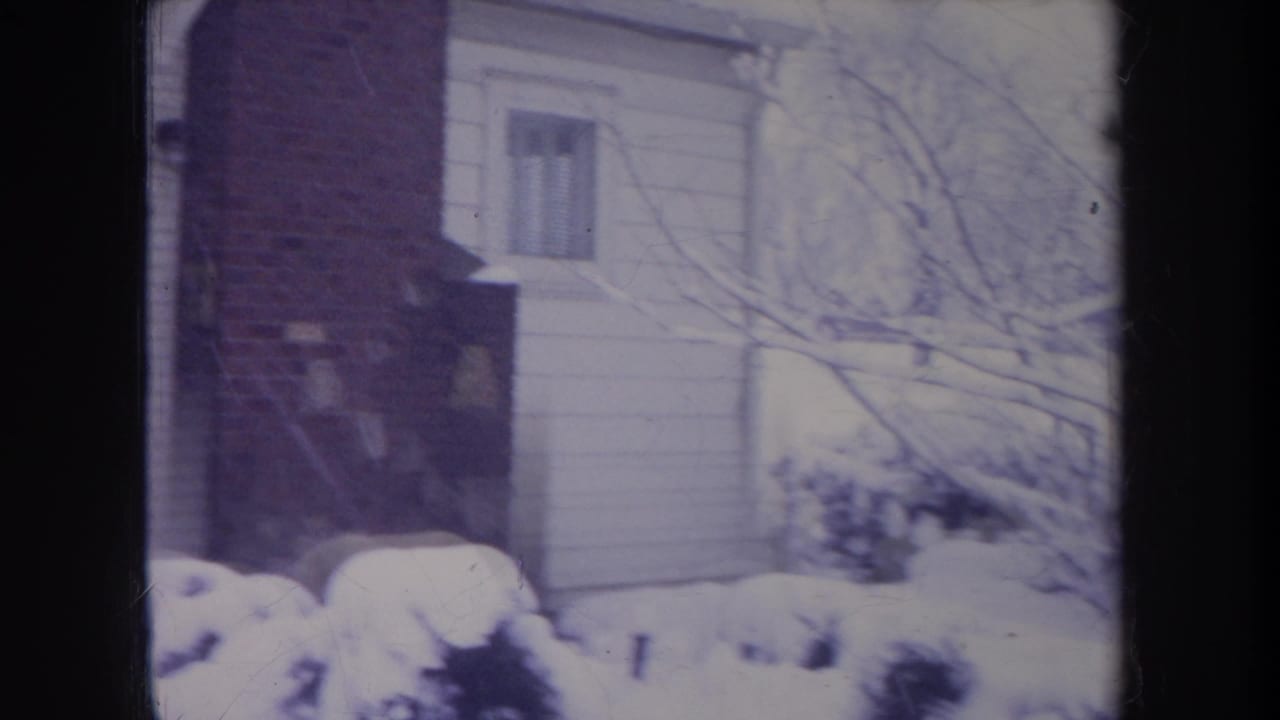 1968 Michigan home-movie frame showing snow on bushes and bare branches beside a white-sided suburban house with brick detail.