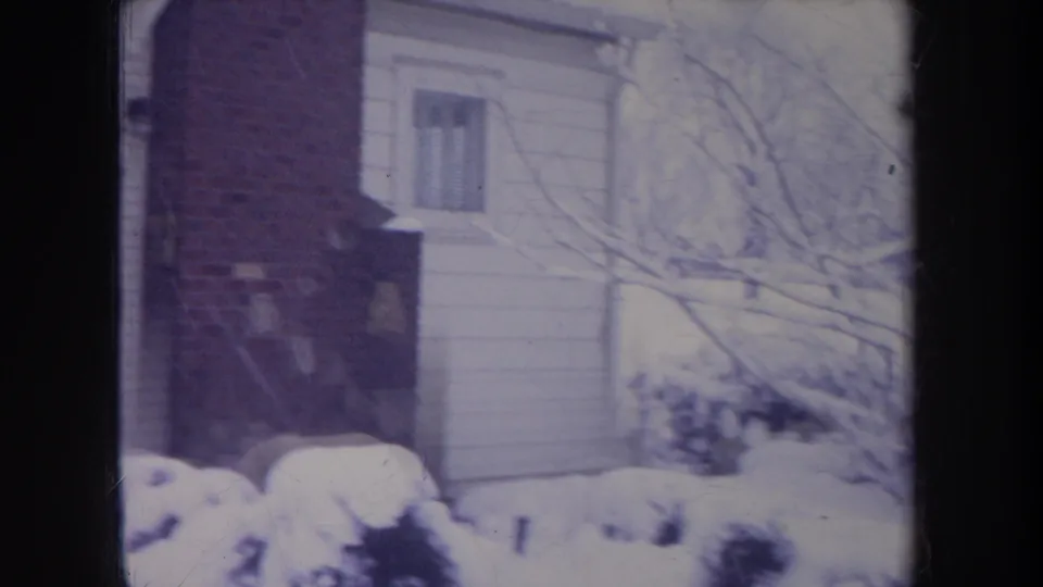 1968 Michigan home-movie frame showing snow on bushes and bare branches beside a white-sided suburban house with brick detail.