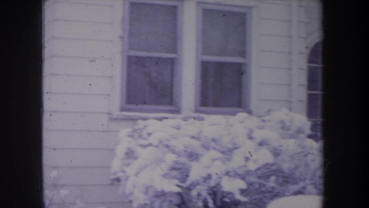 1968 Michigan exterior frame of a white-sided suburban house, two windows, and snow-covered bushes in winter light.