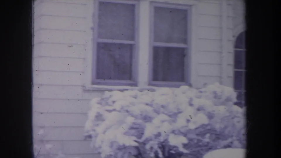 1968 Michigan exterior frame of a white-sided suburban house, two windows, and snow-covered bushes in winter light.