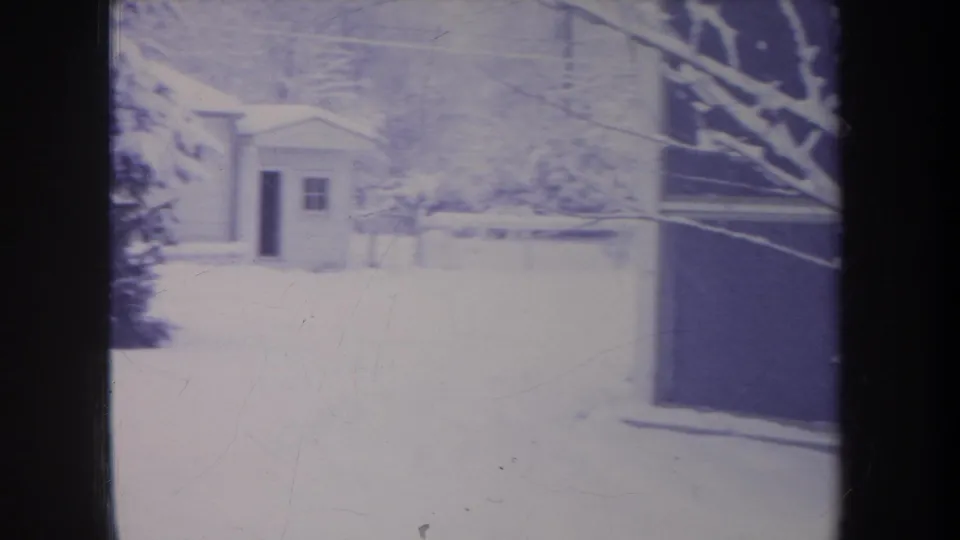 1968 Michigan home-movie frame of a bright snow-covered yard with a small shed or outbuilding beyond trees.