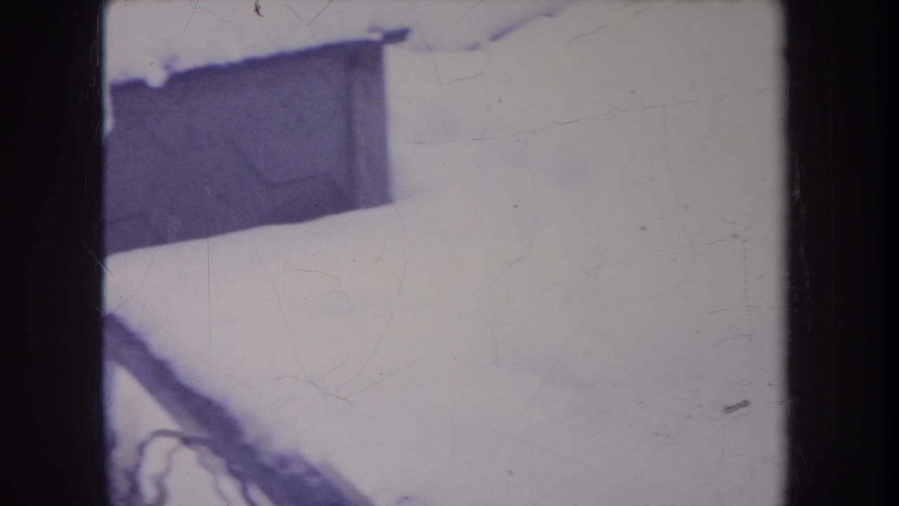Snowy backyard detail with a dark structure and fence edge in faded 1968 Michigan 8mm footage.