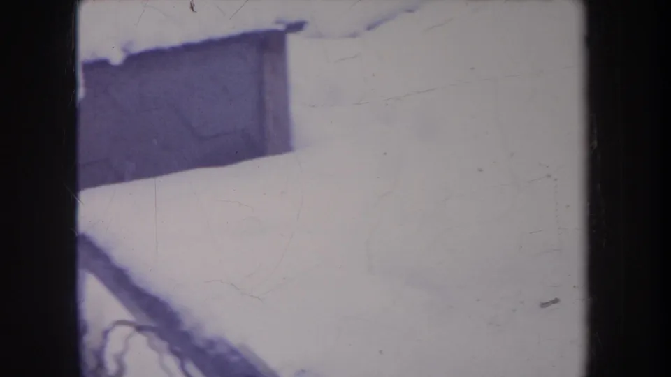 Snowy backyard detail with a dark structure and fence edge in faded 1968 Michigan 8mm footage.