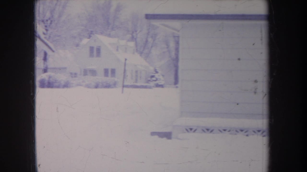 1968 Michigan 8mm frame showing a snow-covered residential yard, neighboring houses, bare trees, and the side of a garage or house.