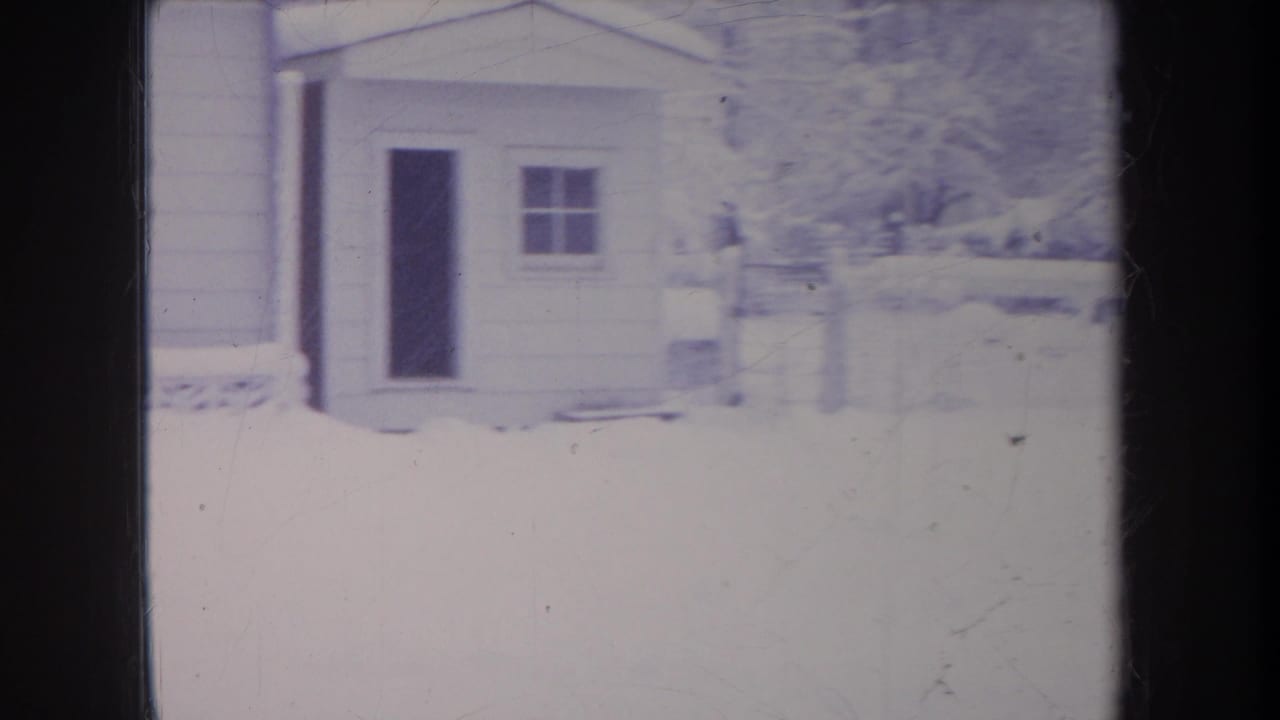 1968 Michigan home-movie frame showing a small outbuilding or side structure with a dark doorway, window, and snow-covered ground.