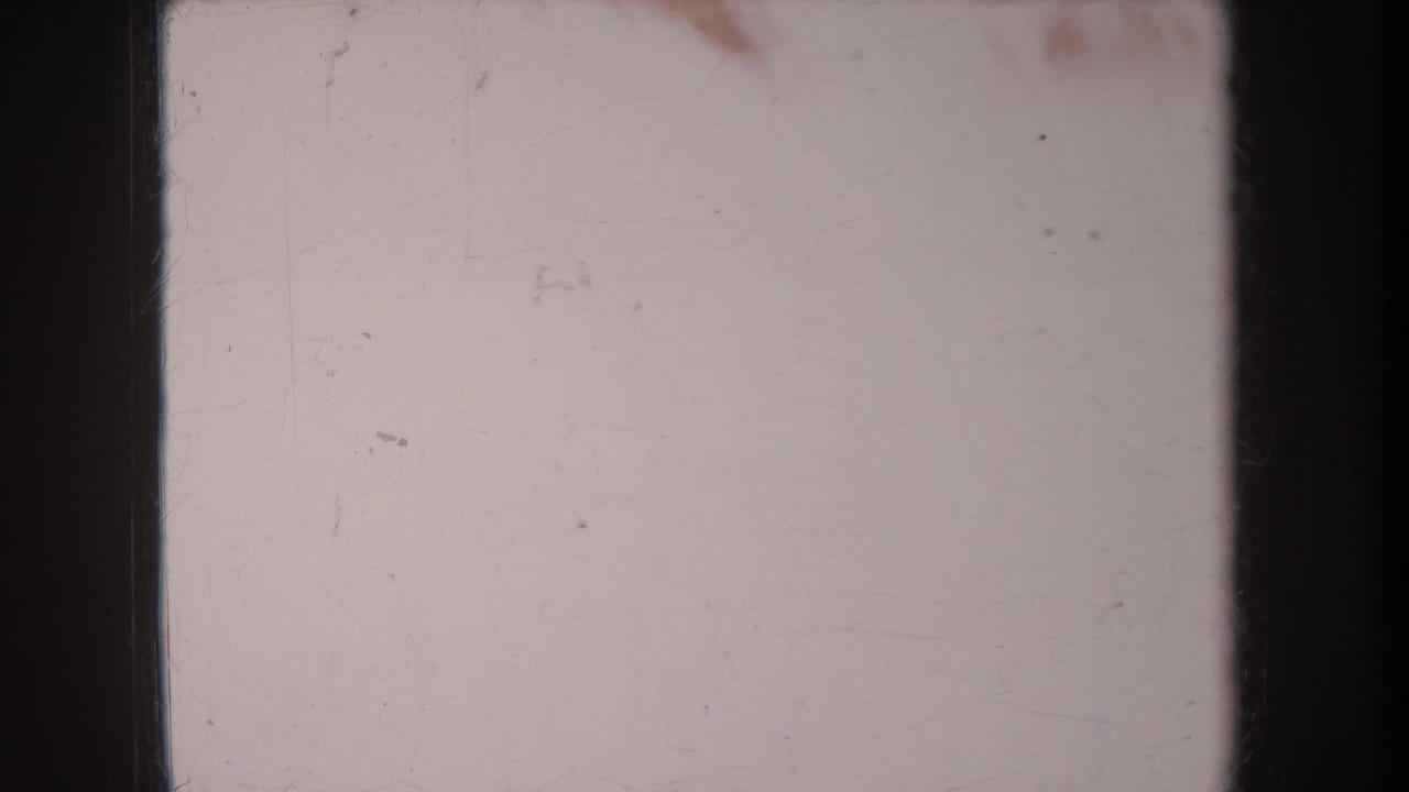 Pale exposed 8mm frame with dark film borders, dust, scratches, and minimal visible scene detail.
