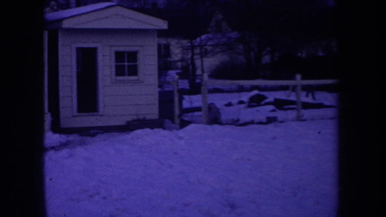 1968 Michigan home-movie frame showing a purple-tinted snowy yard with a small shed, low fence, trees, and frame edge.