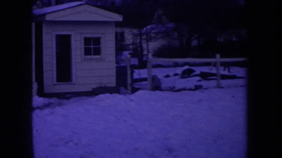 1968 Michigan home-movie frame showing a purple-tinted snowy yard with a small shed, low fence, trees, and frame edge.