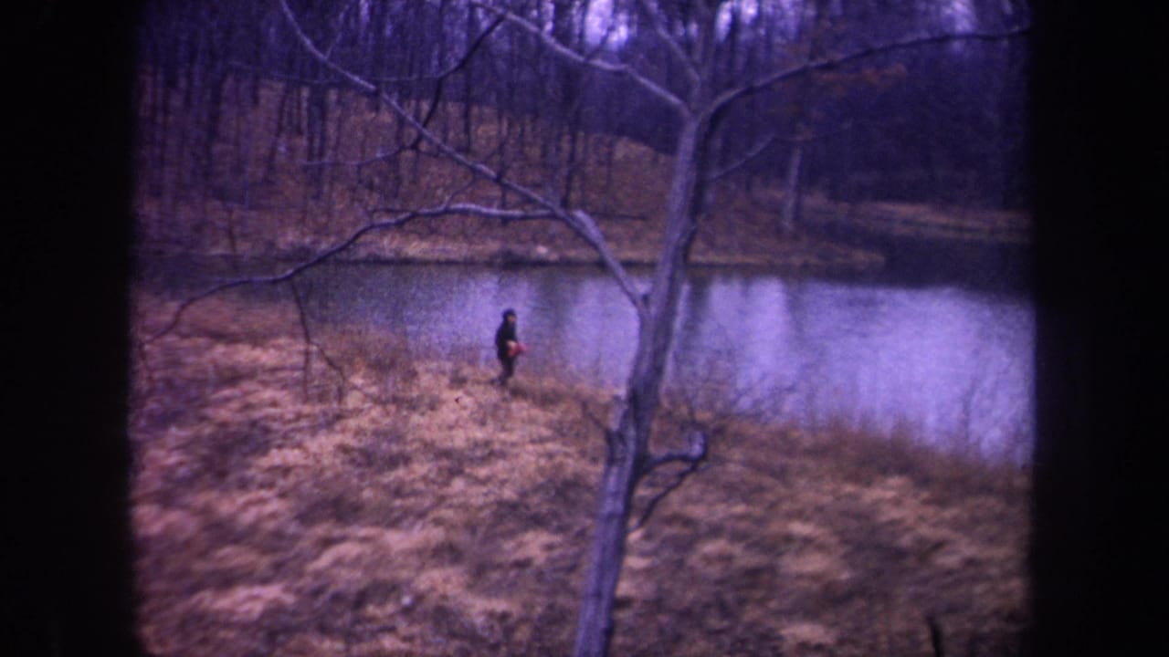 1968 Michigan 8mm frame showing a dark-clothed runner crossing the dry grassy bank beside reflective pond water.