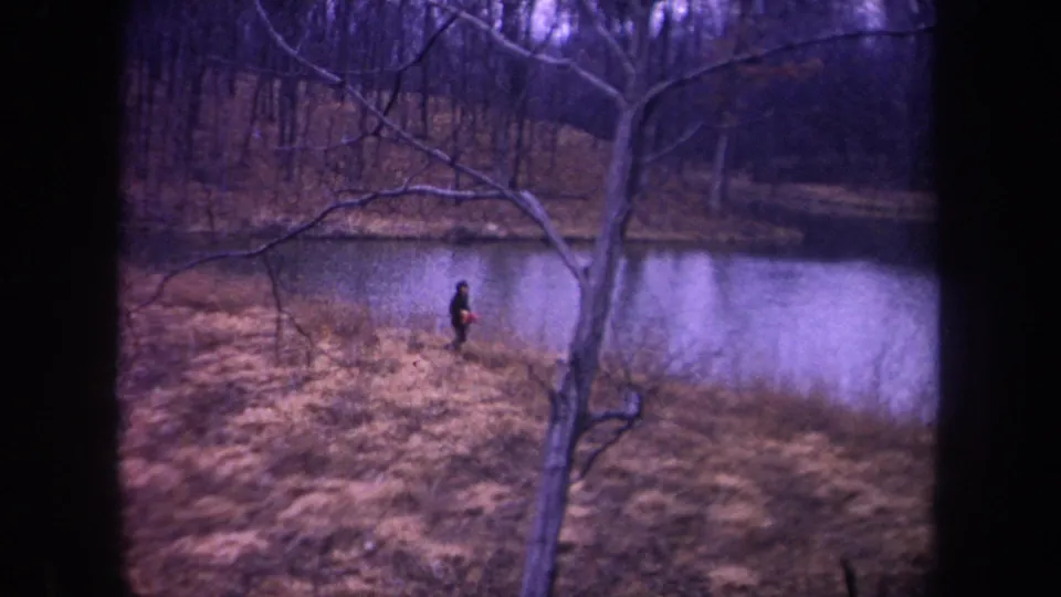 1968 Michigan 8mm frame showing a dark-clothed runner crossing the dry grassy bank beside reflective pond water.
