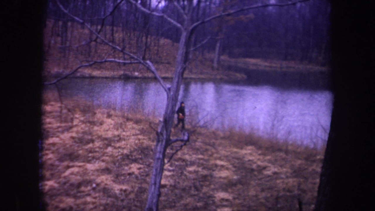 1968 Michigan home-movie frame of a man running near a foreground tree, with pond water and bare woodland behind him.