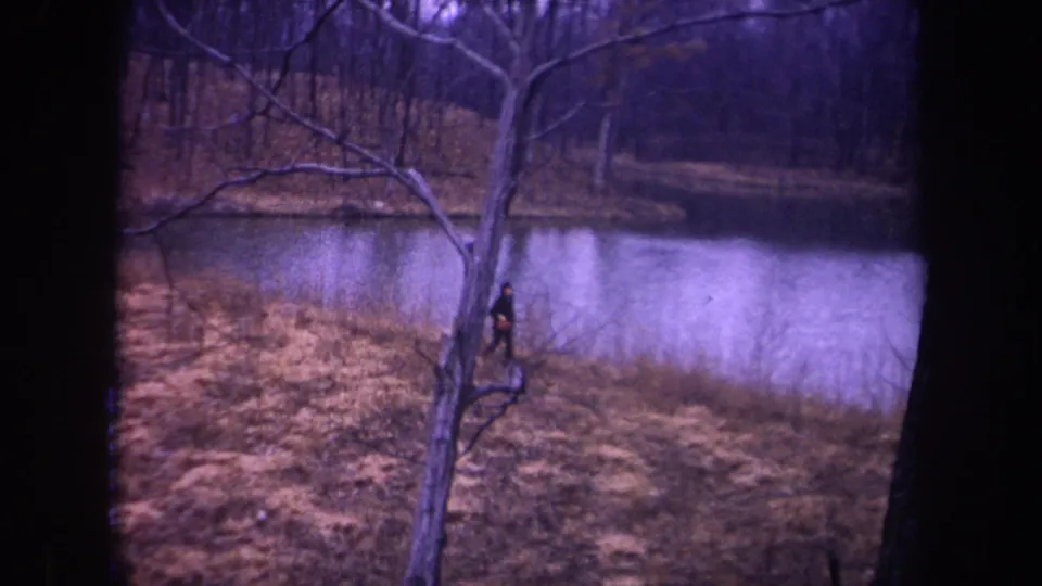1968 Michigan home-movie frame of a man running near a foreground tree, with pond water and bare woodland behind him.