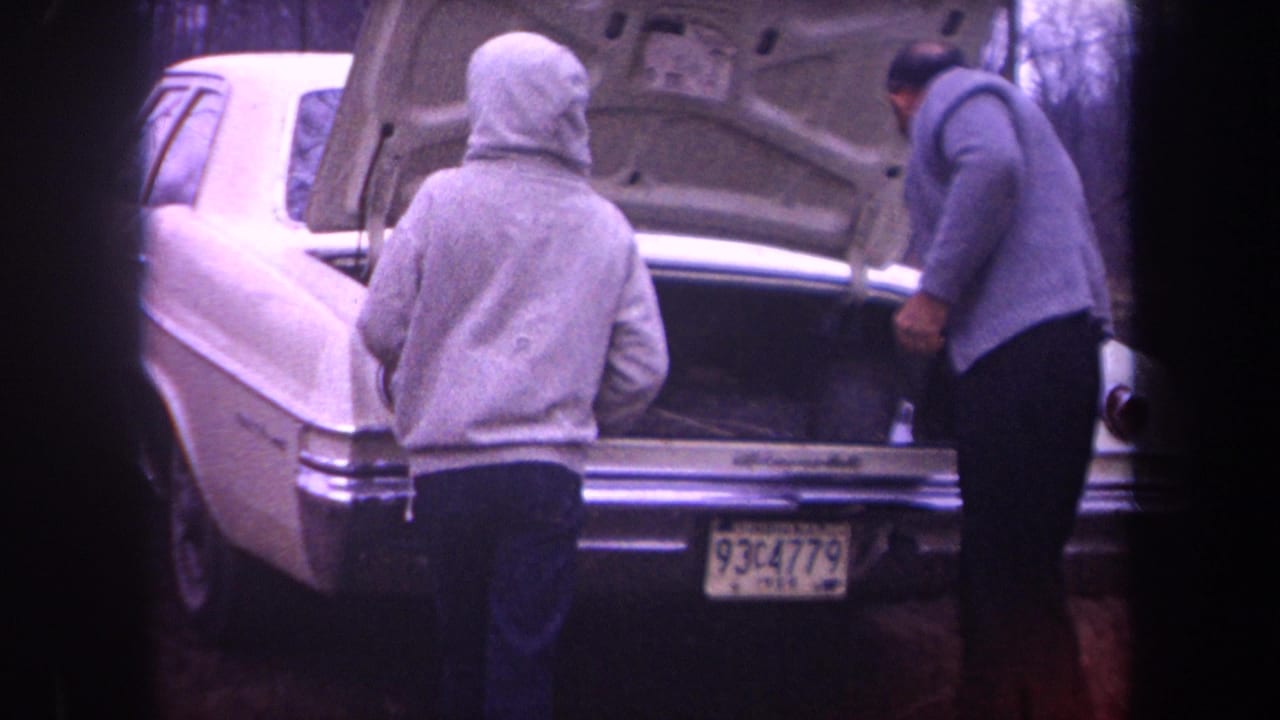 Two men stand behind a white sedan with its trunk open in a wooded area.