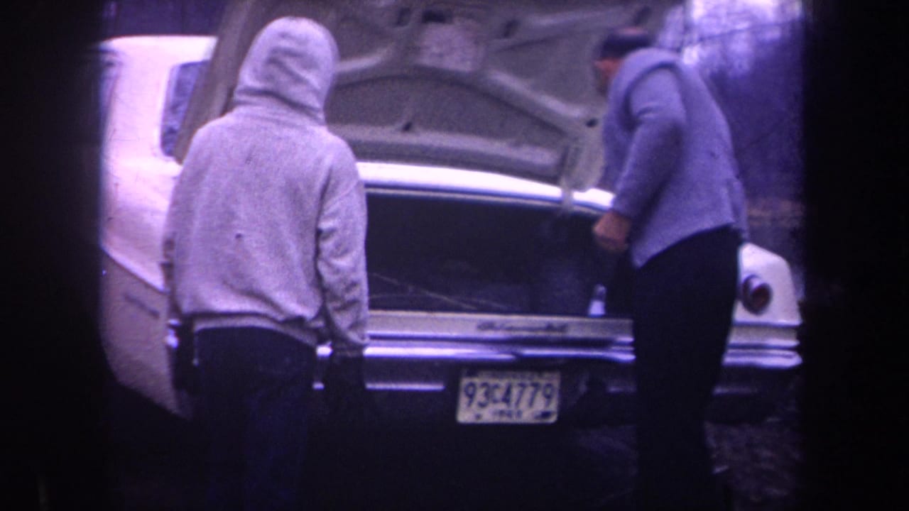 A hooded man and an older man stand at the open trunk of a white car.