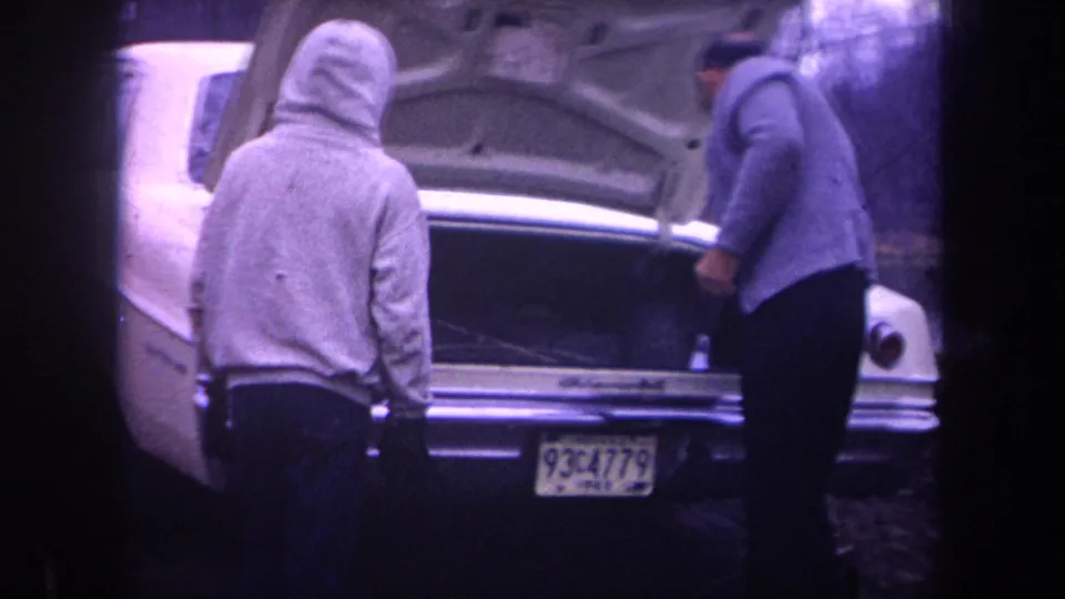 1968 Michigan home-movie frame with a hooded man and older man beside the open trunk of a white sedan.