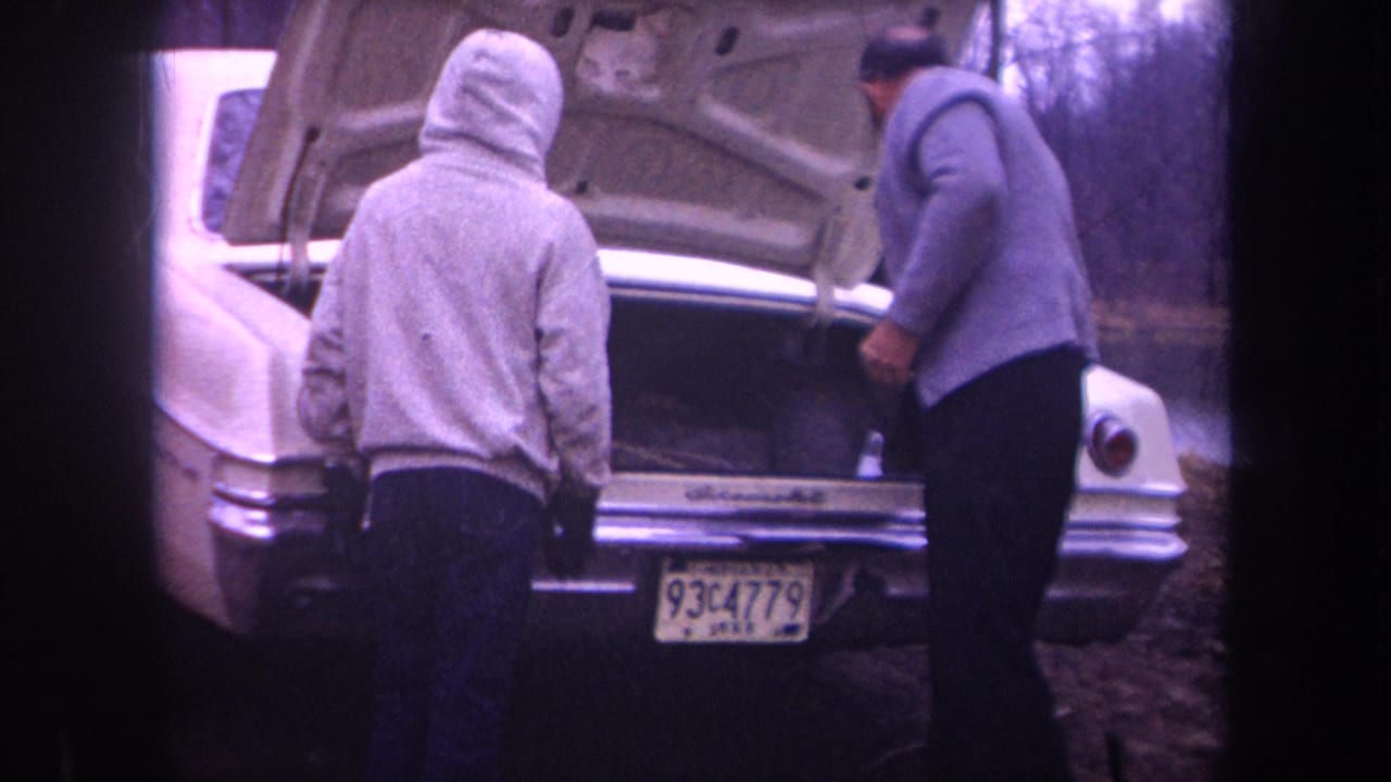 Soft 8mm frame from Michigan showing two men at the rear of a white sedan with the trunk lid raised.