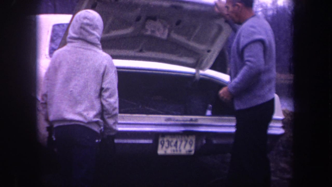 1968 Michigan frame showing an older man in a sweater and cap beside a hooded man at the open trunk of a white sedan.
