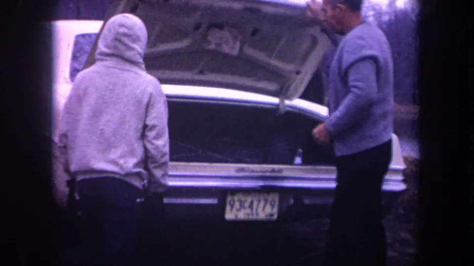 1968 Michigan frame showing an older man in a sweater and cap beside a hooded man at the open trunk of a white sedan.