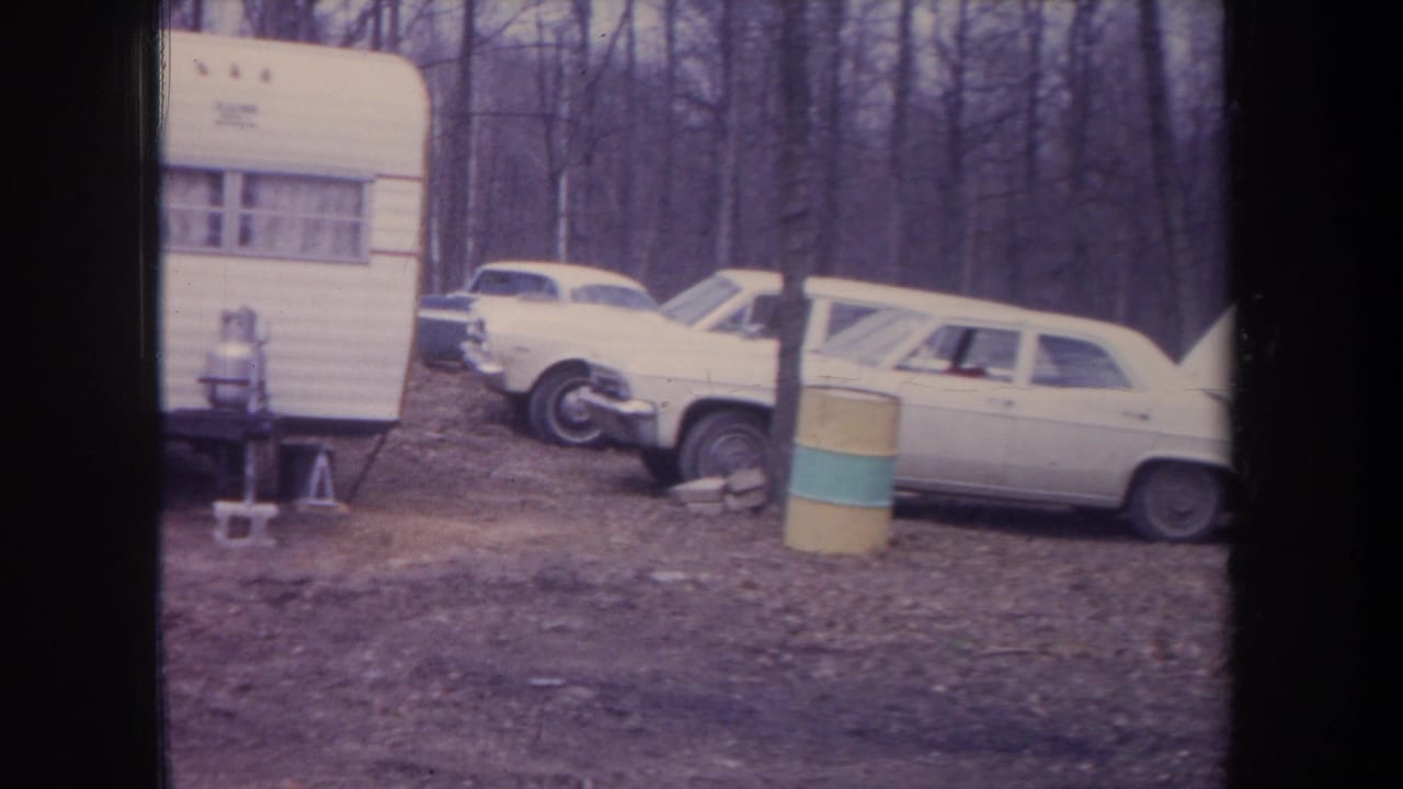 1968 Michigan home-movie frame showing a white camper, classic cars, striped barrel, and wooded autumn campsite.