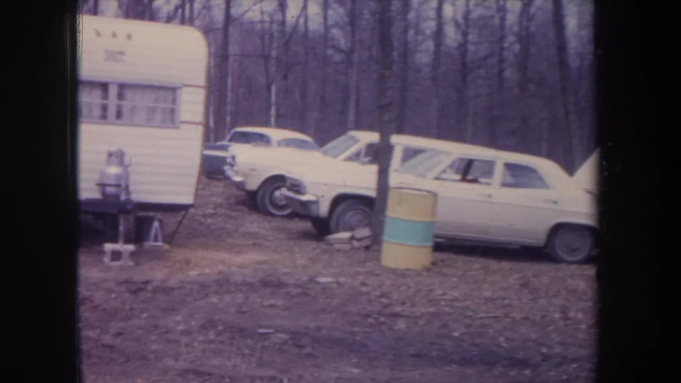 1968 Michigan home-movie frame showing a white camper, classic cars, striped barrel, and wooded autumn campsite.
