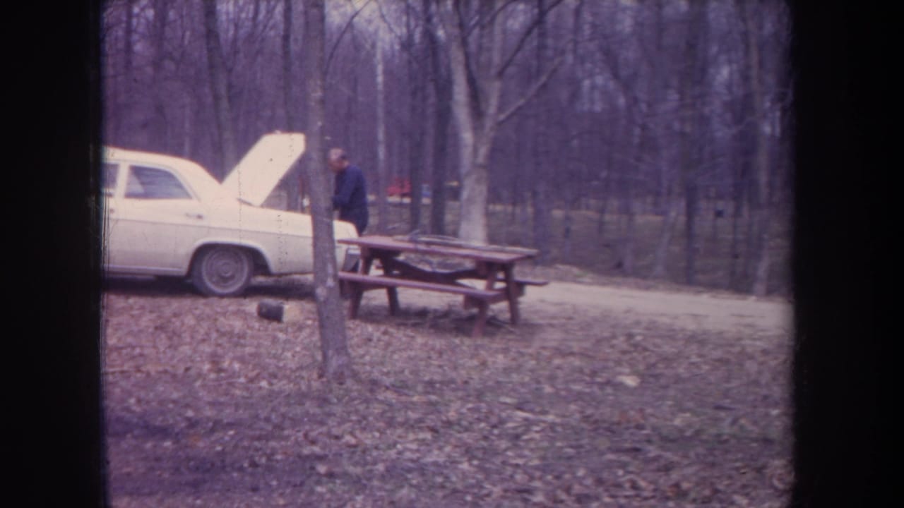 1968 Michigan campsite view with an adult near a picnic table, a white sedan with raised hood, and bare autumn trees.
