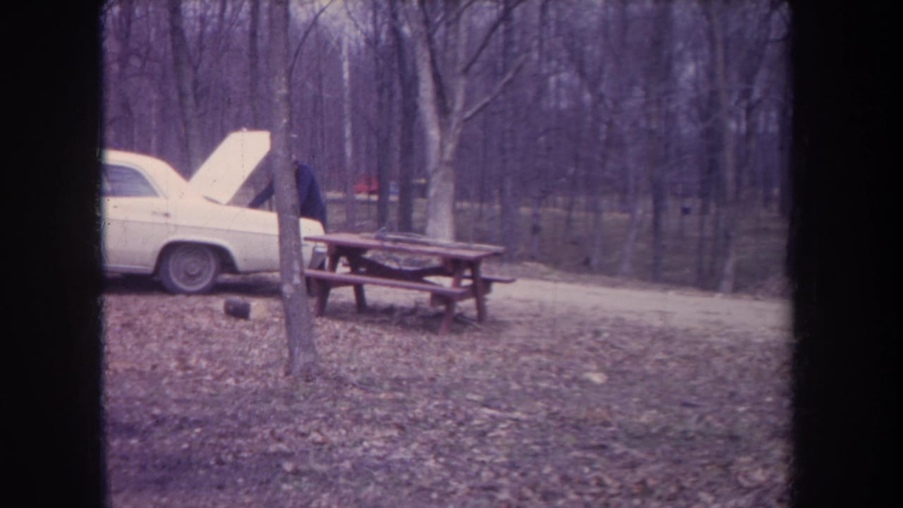 1968 Michigan home-movie frame showing a white car with raised hood, adult figure, picnic table, and leaf-covered woods.