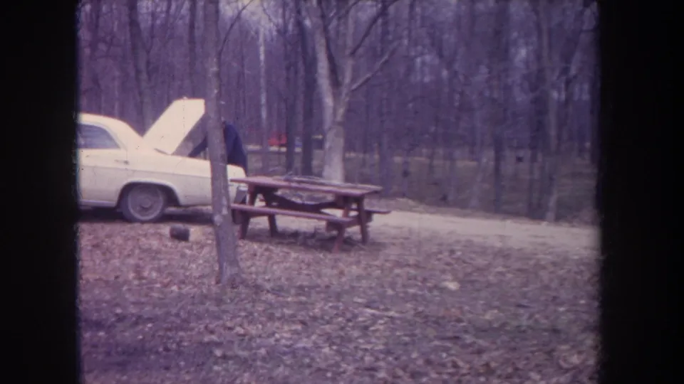 1968 Michigan home-movie frame showing a white car with raised hood, adult figure, picnic table, and leaf-covered woods.