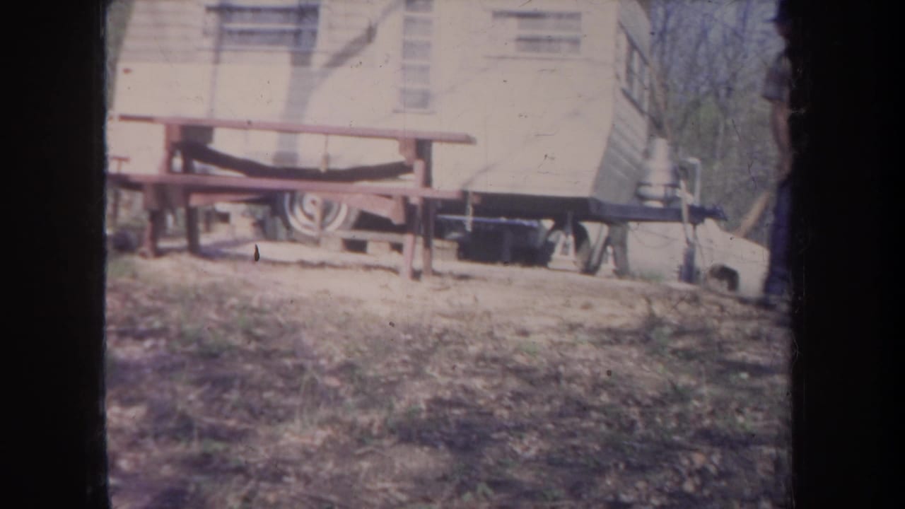 1968 Michigan 8mm frame showing a light-colored trailer behind a wooden picnic table in a leaf-covered wooded campsite.