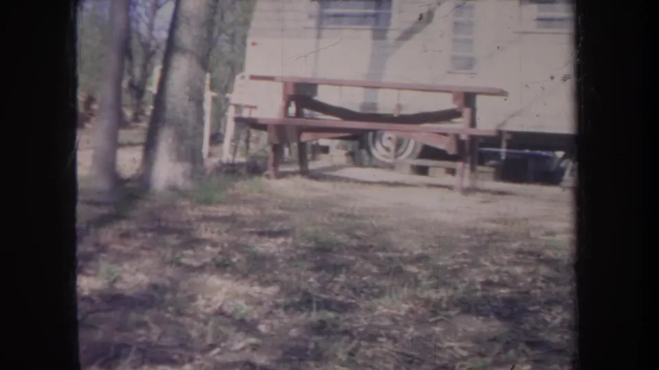 1968 Michigan home-movie frame with a trailer, picnic table, trees, and bare ground in a wooded campsite clearing.