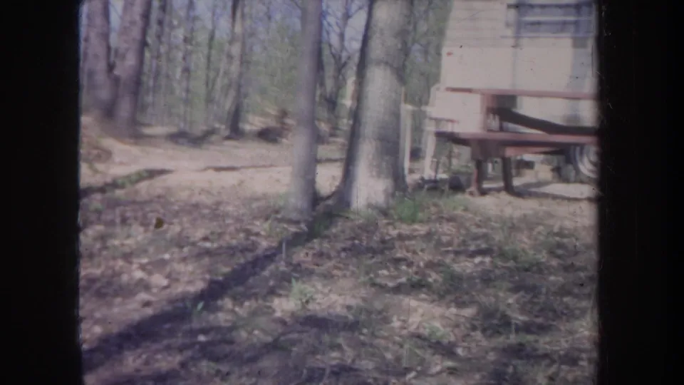 1968 Michigan 8mm frame showing the edge of a trailer, trees, and uneven leaf-covered ground in a wooded campsite.
