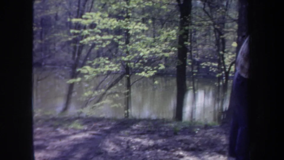 1968 Michigan 8mm frame showing a wooded riverside area, reflective water, and a partially obscured man entering the scene.