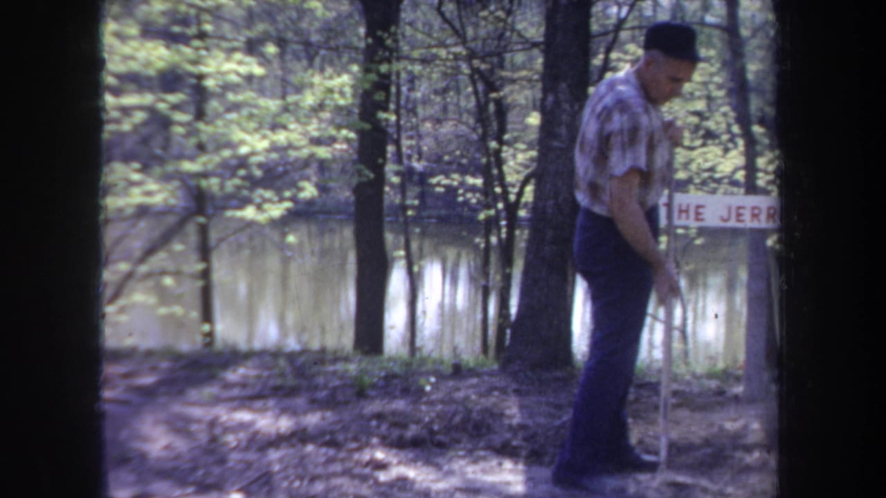 Man in plaid shirt stands by a riverside sign with water behind him.