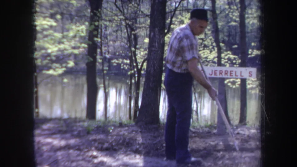 1968 Michigan home-movie frame of a man working with a long-handled tool near a Jerrell's sign on a wooded shoreline.