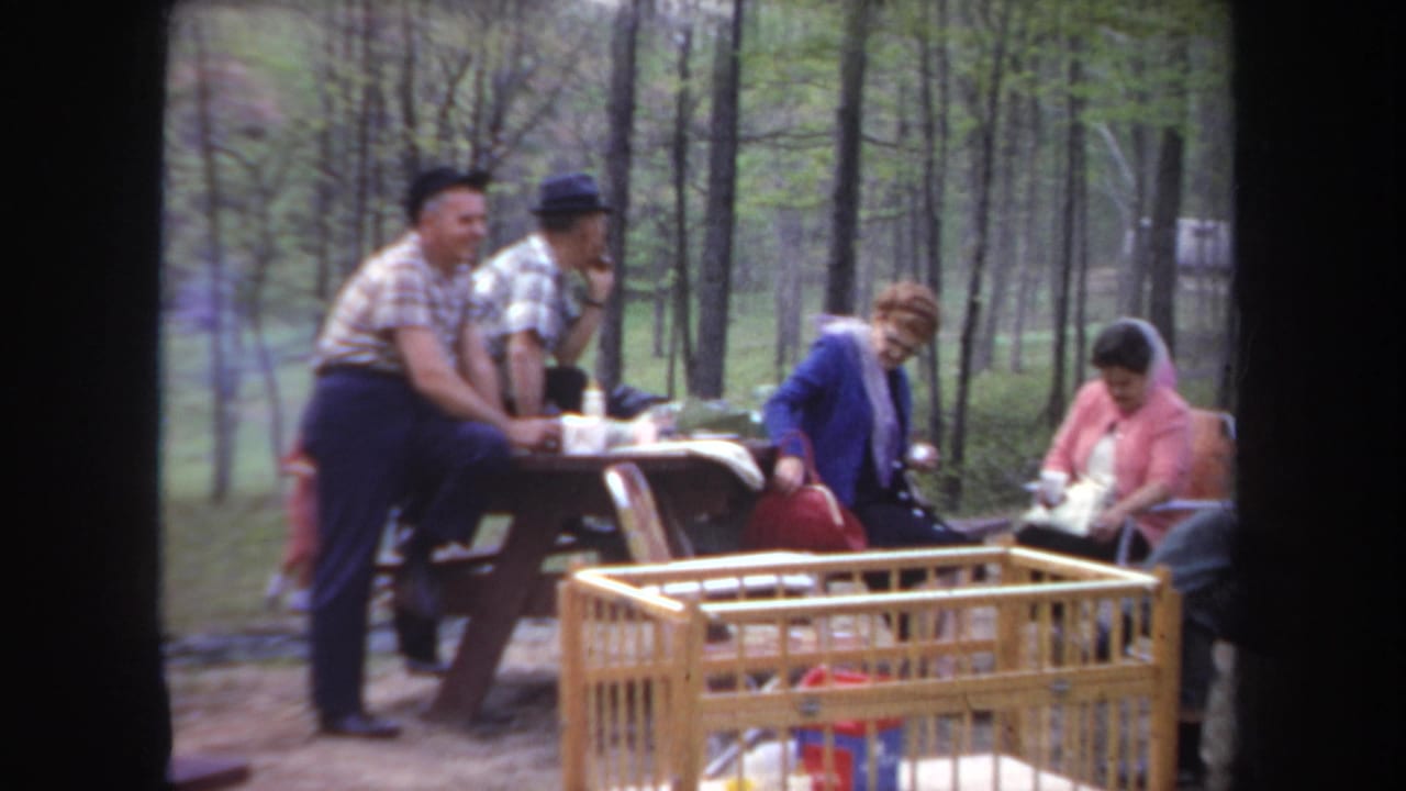 1968 Michigan home-movie frame showing adults near a picnic table in a wooded campground, with a wooden baby crib in the foreground.
