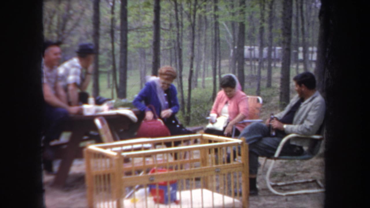 Adults sit and stand near a campsite picnic table, folding chairs, and a crib among trees.