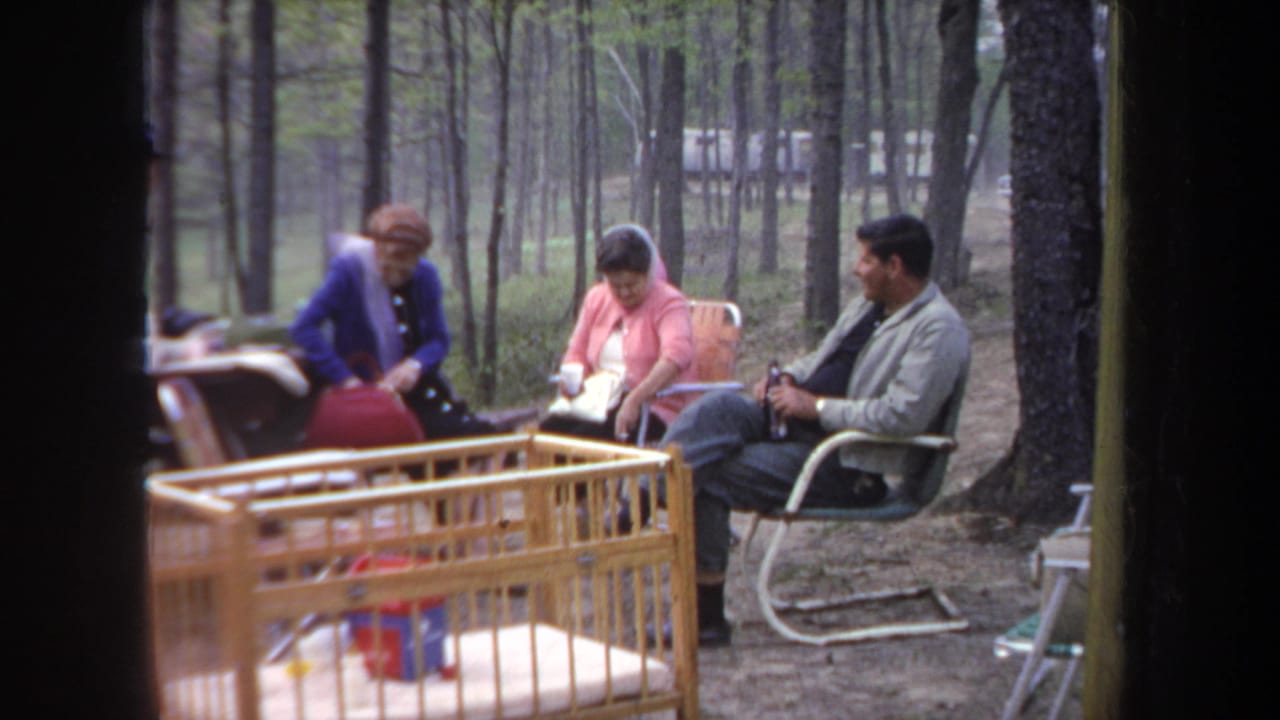 1968 Michigan home-movie frame of seated adults at a wooded campsite, with a picnic table edge and wooden crib in front.