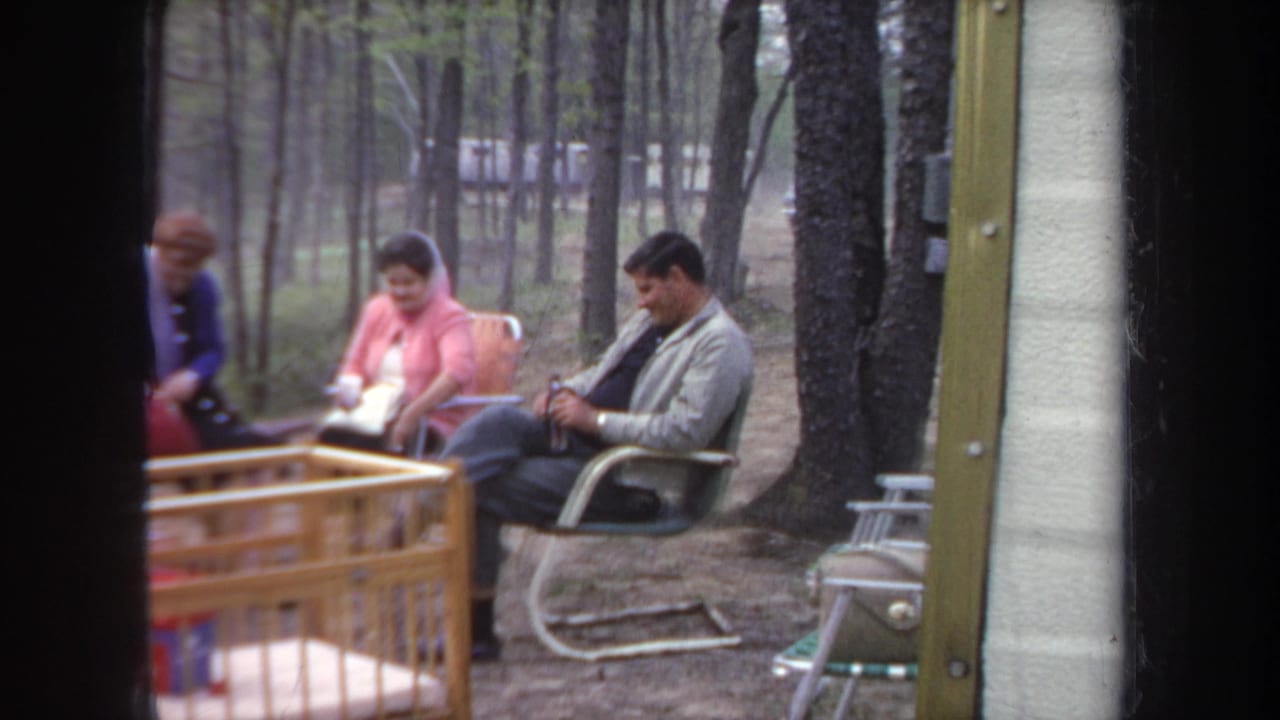 1968 Michigan campsite frame showing two seated adults near a tree, folding chairs, a crib edge, and camping items.