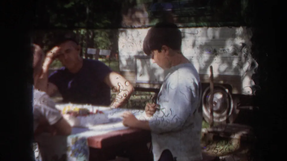 A candid 1968 Michigan home-movie frame shows adults around an outdoor table with trees and a light-colored trailer or vehicle behind them.