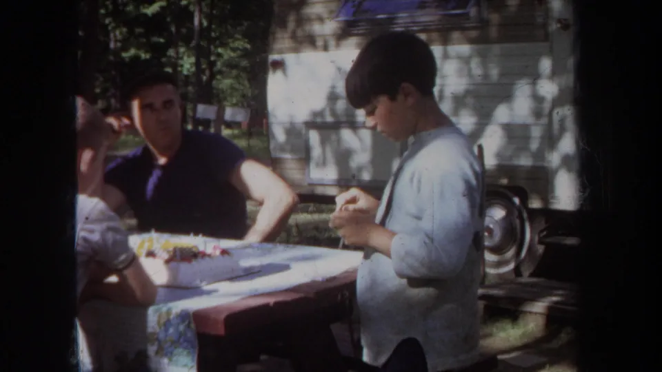A person stands beside a family table while seated adults remain nearby in a shaded Michigan outdoor home-movie scene from 1968.