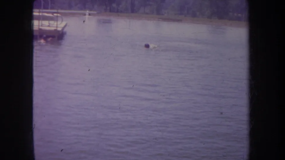 A swimmer moves through a Michigan lake or river near a small boat or dock in 1968 8mm footage.