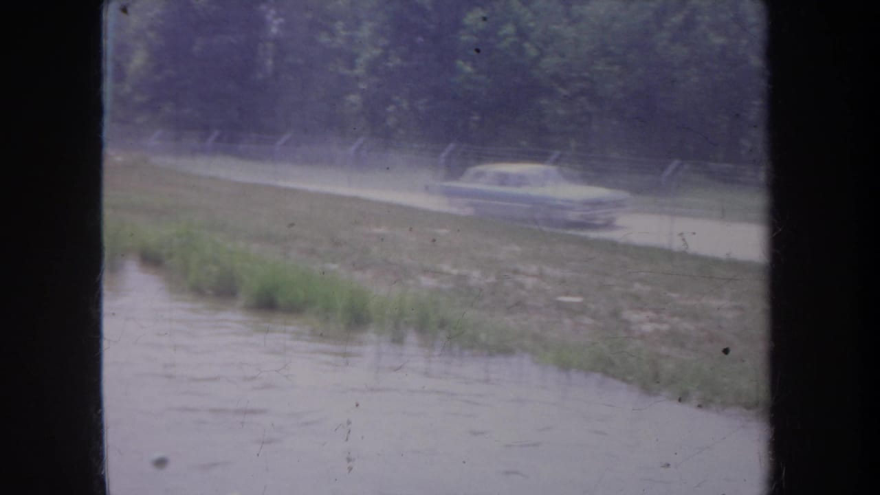 1968 Michigan home-movie frame showing a classic sedan on a wet rural road beside floodwater, viewed through dark window mullions.