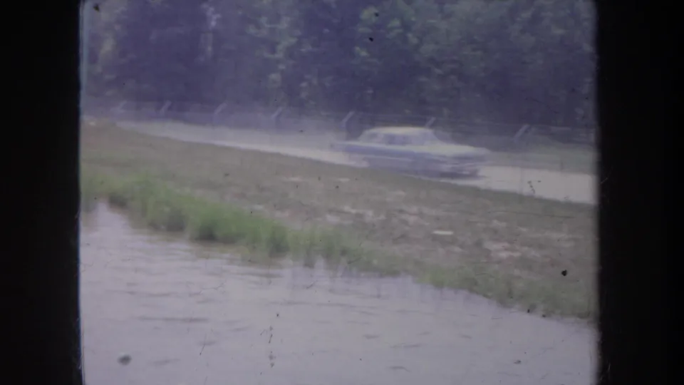 1968 Michigan home-movie frame showing a classic sedan on a wet rural road beside floodwater, viewed through dark window mullions.